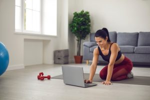 Happy fit woman sitting on sports mat on the floor at home and using laptop computer. Smiling lady watching video fitness workout lesson or shopping for sportswear and sporting equipment online